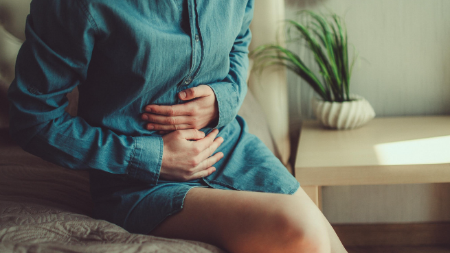 Une femme en chemise bleu lagon assise sur un lit se tient le ventre avec les mains. Une plante verte est dans le fond sur une petite table